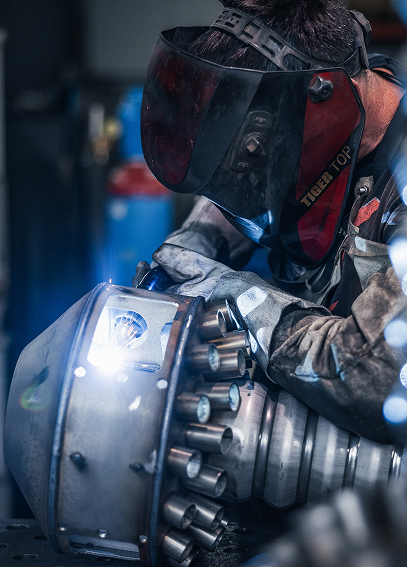 Ein Mann mit Schweißhelm und Schutzausrüstung arbeitet an einem Metallteil, aus dem Funken sprühen, in einer Werkstatt.