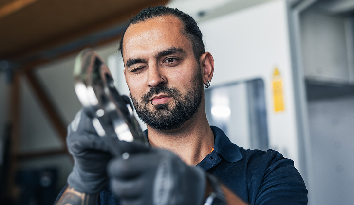 Ein Mann mit Bart untersucht ein glänzendes Metallteil. Er trägt schwarze Handschuhe und kneift ein Auge zusammen.