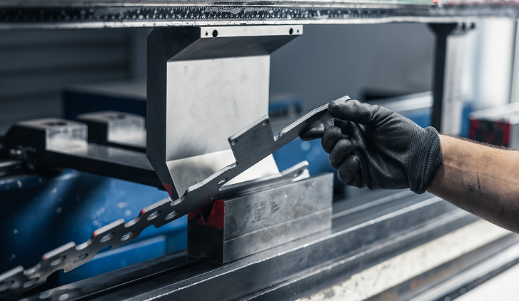 Eine behandschuhte Hand hält ein gebogenes Metallstück vor einer Biegemaschine in einer Werkstatt.