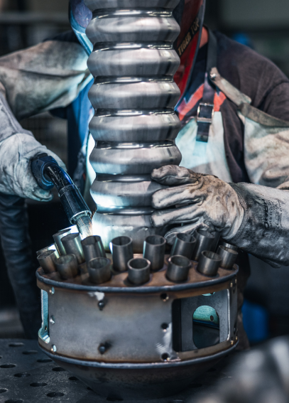 Ein Schweißer arbeitet an einem großen Metallteil mit mehreren Rohren. Er trägt Schutzkleidung und benutzt ein Schweißgerät.