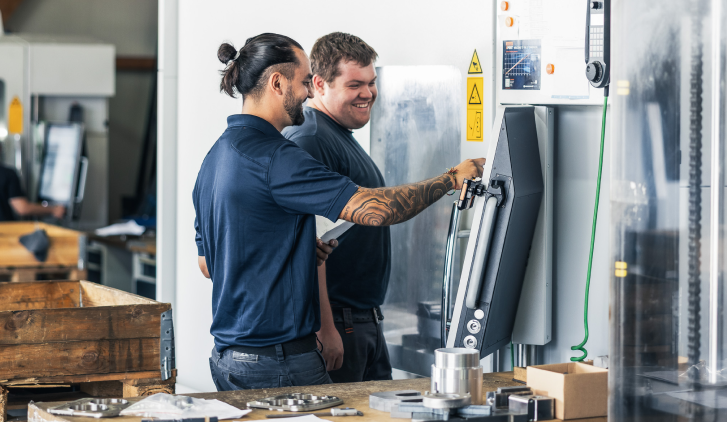 Zwei Männer in Arbeitskleidung bedienen eine CNC-Maschine in einer Werkstatt. Einer zeigt lächelnd auf den Bedienbildschirm.