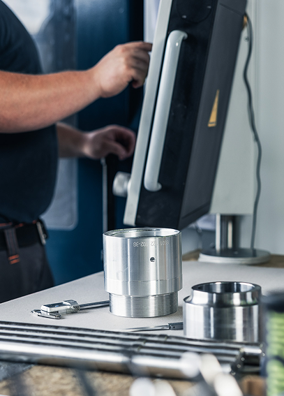 Eine Person bedient einen Computer in einer Werkstatt. Im Vordergrund sind metallische Werkstücke und ein Messschieber zu sehen.