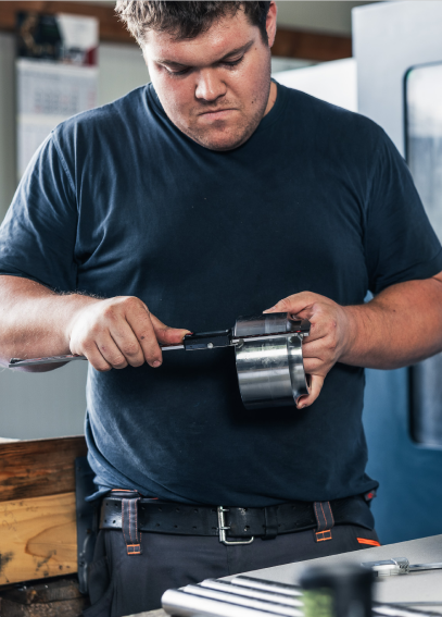 Ein Mann in Arbeitskleidung misst ein Metallteil mit einem Messschieber in einer Werkstattumgebung.