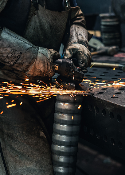 Ein Arbeiter schleift an einem Metallwerkstück, wobei Funken sprühen. Er trägt Schutzkleidung, einschließlich Handschuhe und Schürze.