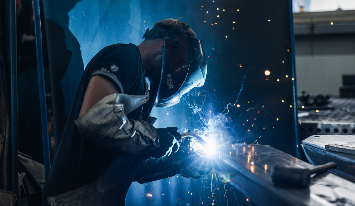 Ein Schweißer mit Schutzhelm und Handschuhen arbeitet an einem Metallstück, während Funken in einer Werkstatt fliegen.
